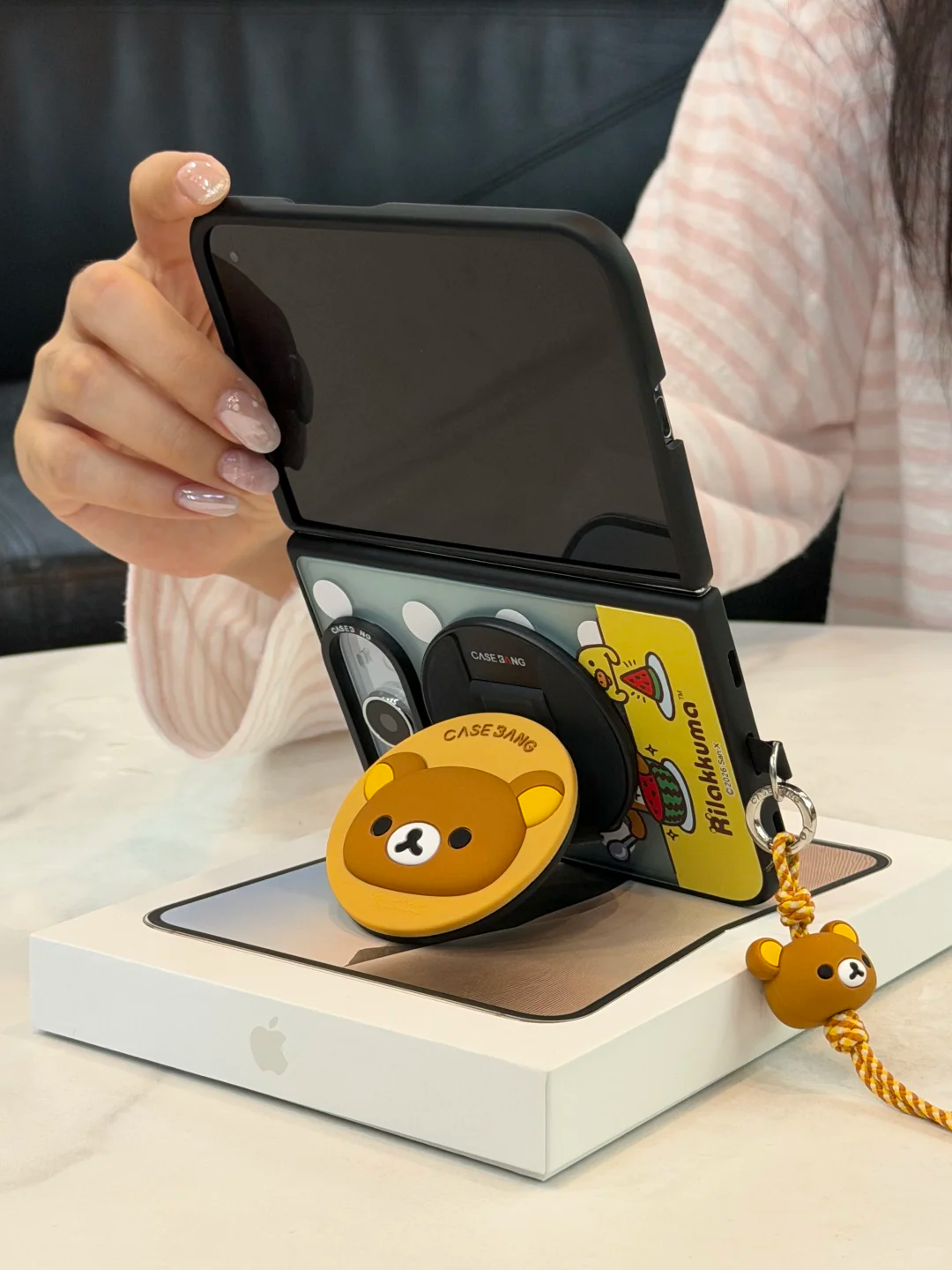 Original real photograph of a foldable phone in a case with a Rilakkuma round stand and lanyard. A hand holds the upper screen.